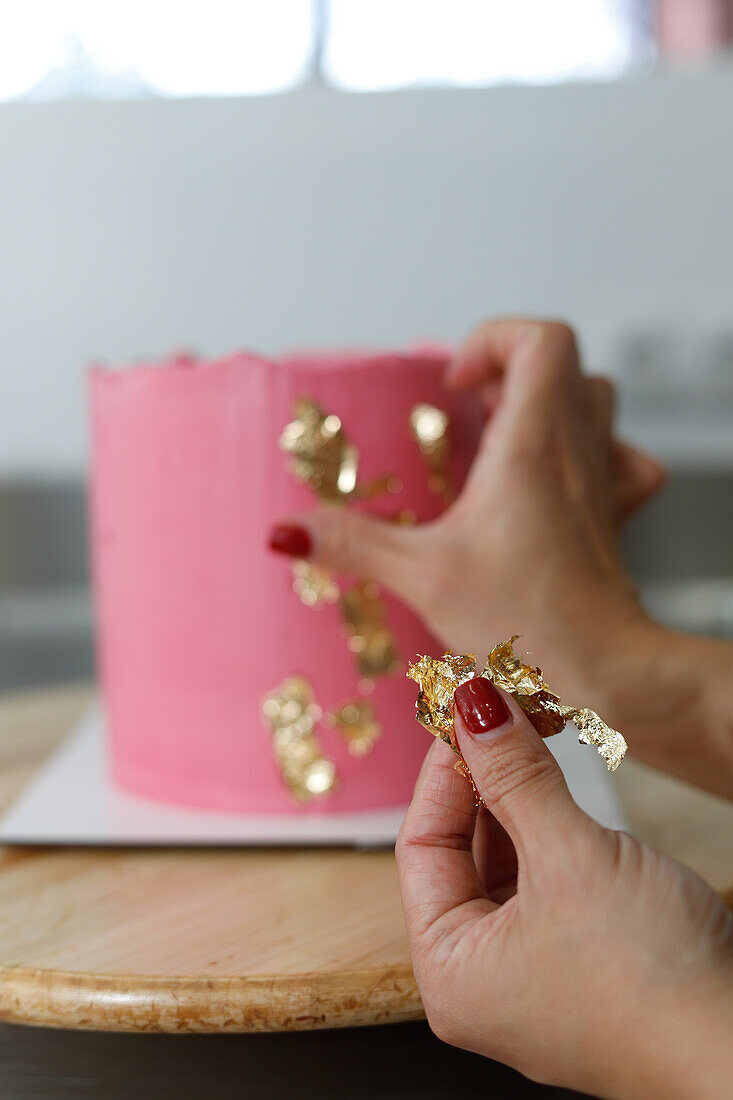 Close-up of a person applying delicate gold leaf to a vibrantly pink frosted cake, highlighting the art of cake decoration