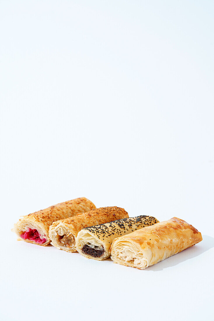 A selection of flaky pastry rolls with various fillings, garnished with seeds, on a clean white background.