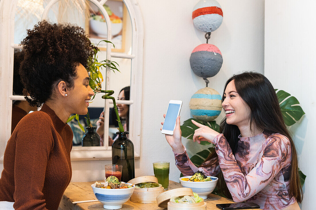 Cheerful diverse female friends sitting at table with poke in restaurant and using smartphone while spending weekend together