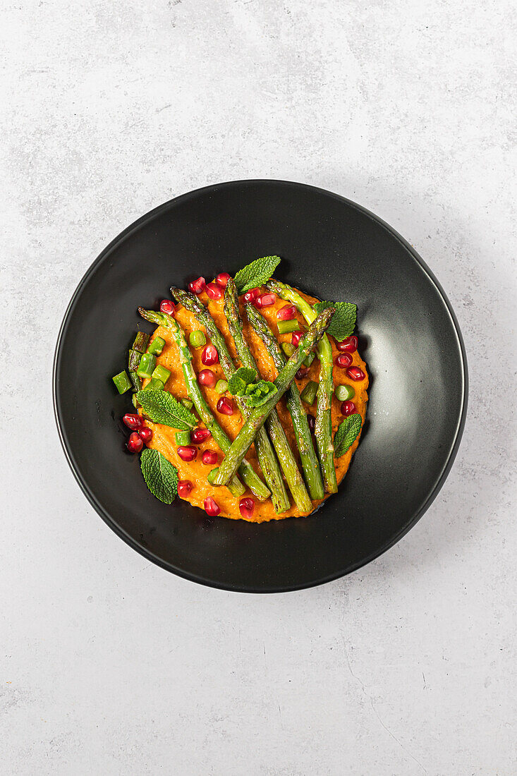 Top view image of a wholesome dish with grilled asparagus and pomegranate seeds on a bed of mashed sweet potato garnished with mint leaves, served on a black plate against a grey background.