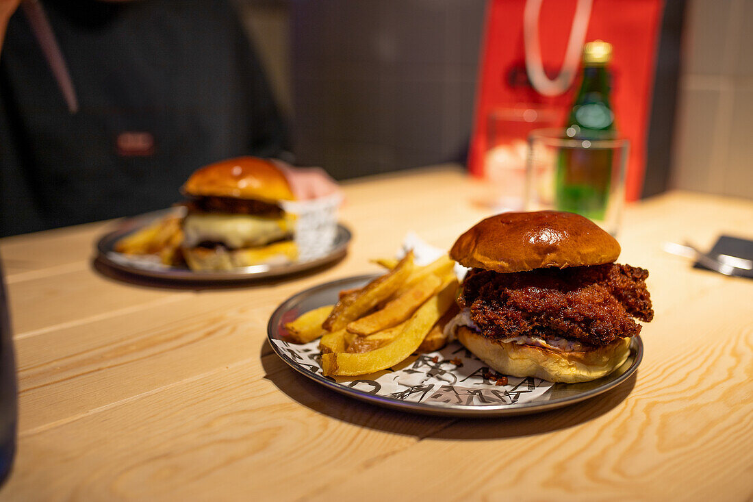 Two gourmet burgers with a side of crispy French fries on patterned ceramic plates A soft drink and blurred background enhance the cozy dining setting