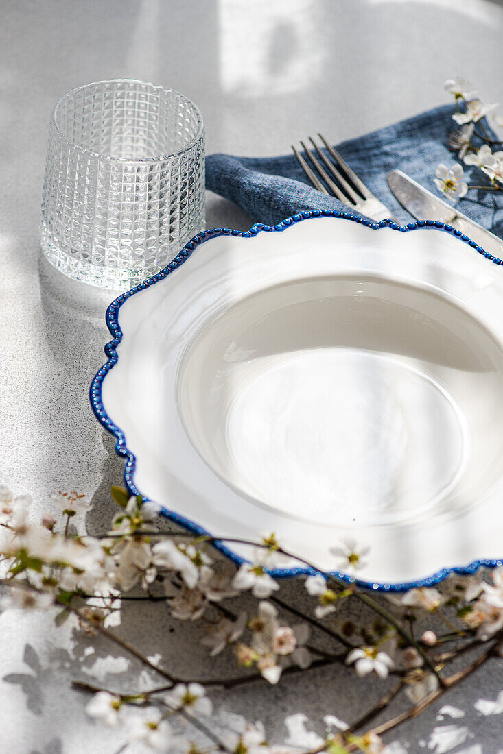 A sophisticated table setting featuring a white plate with blue detailing, glassware, and a sprig of spring blossoms on a textured surface