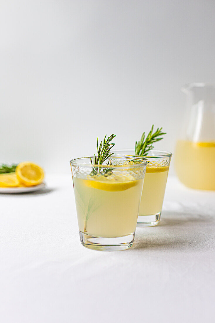 Two glasses of freshly made lemonade with rosemary sprigs and a sliced lemon in the background on a clean white surface