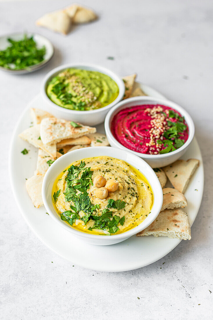 From above image showcases a selection of three hummus varieties - classic, beet, and avocado with spinach - garnished with fresh herbs, accompanied by pita bread on a white plate.