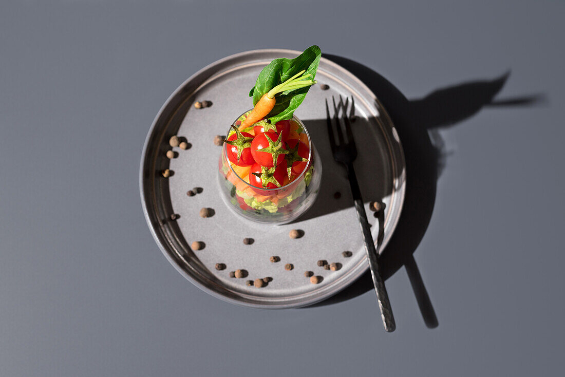 Top view of a colorful vegetable salad neatly presented in a transparent glass, placed on a textured gray plate. The arrangement includes fresh greens, cherry tomatoes, and a carrot, accompanied by a fork and scattered chickpeas around the plate.