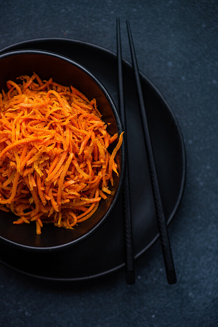 A vertical image capturing a close-up of fresh grated carrot salad served in a stylish black bowl with a pair of black chopsticks on a dark background