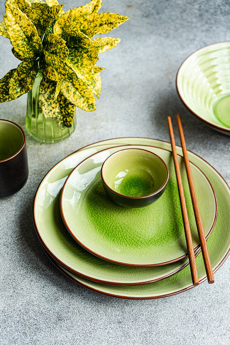 From above view of a modern table setting featuring bright green ceramic dinnerware and wooden chopsticks, juxtaposed with a vibrant potted plant, all showcased on a textured grey background.