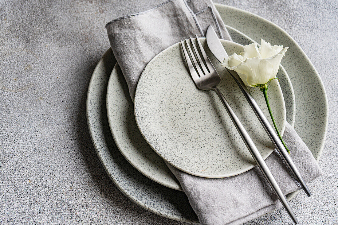 An elegant table setting featuring a stack of speckled ceramic plates, modern silver cutlery, a linen napkin, and a delicate godetia flower, all presented on a textured surface