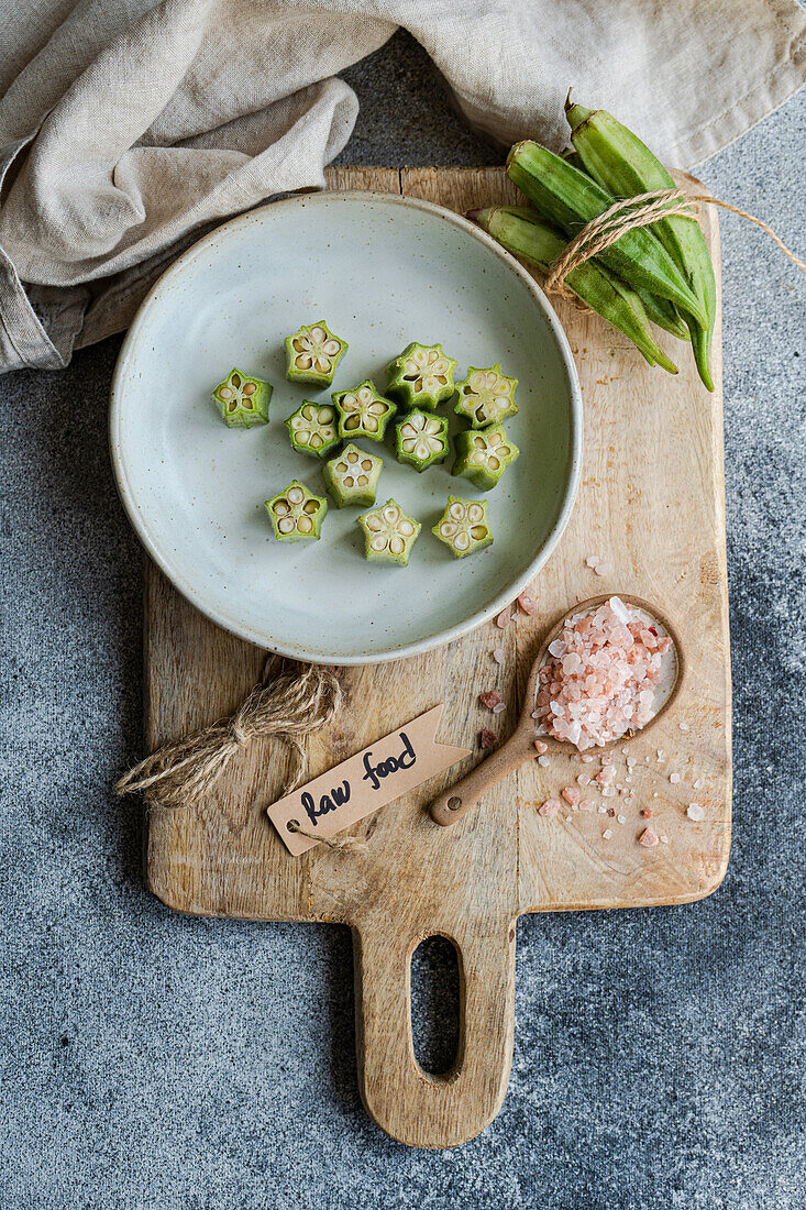 In Scheiben geschnittene Okra-Bamia, ordentlich auf einem Keramikteller angerichtet, präsentiert auf einem rustikalen Holzbrett neben einem Löffel mit rosa Salz, mit einem Rohkost-Holzschild, das eine rohe, natürliche Lebensmittelzubereitung hervorhebt