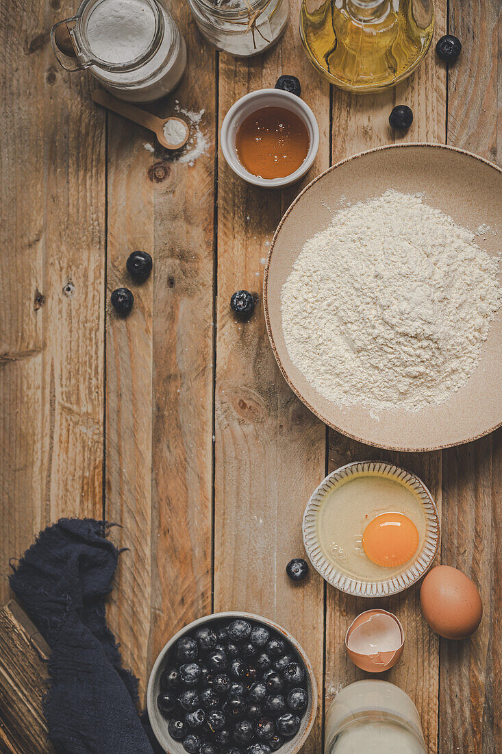 Top view of a wooden table filled with baking essentials such as flour, egg, honey, and fresh blueberries, evoking a cozy, homemade baking atmosphere