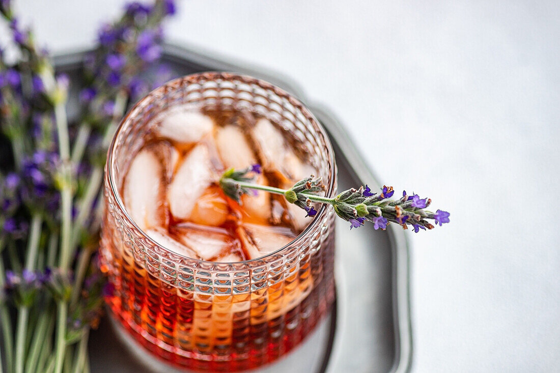 A sophisticated cognac cocktail, served in a textured glass with ice cubes, garnished with a fresh sprig of lavender, displayed on a metal tray with a subtle backdrop