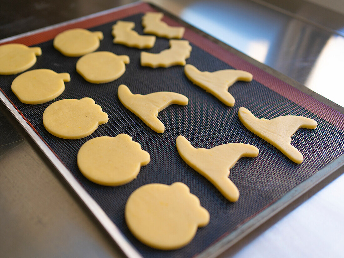 Halloween-Kekse in Form von Fledermäusen, Hexenhüten und Kürbissen auf einem Backblech, bereit zum Backen Ideal für festliche Backaktivitäten