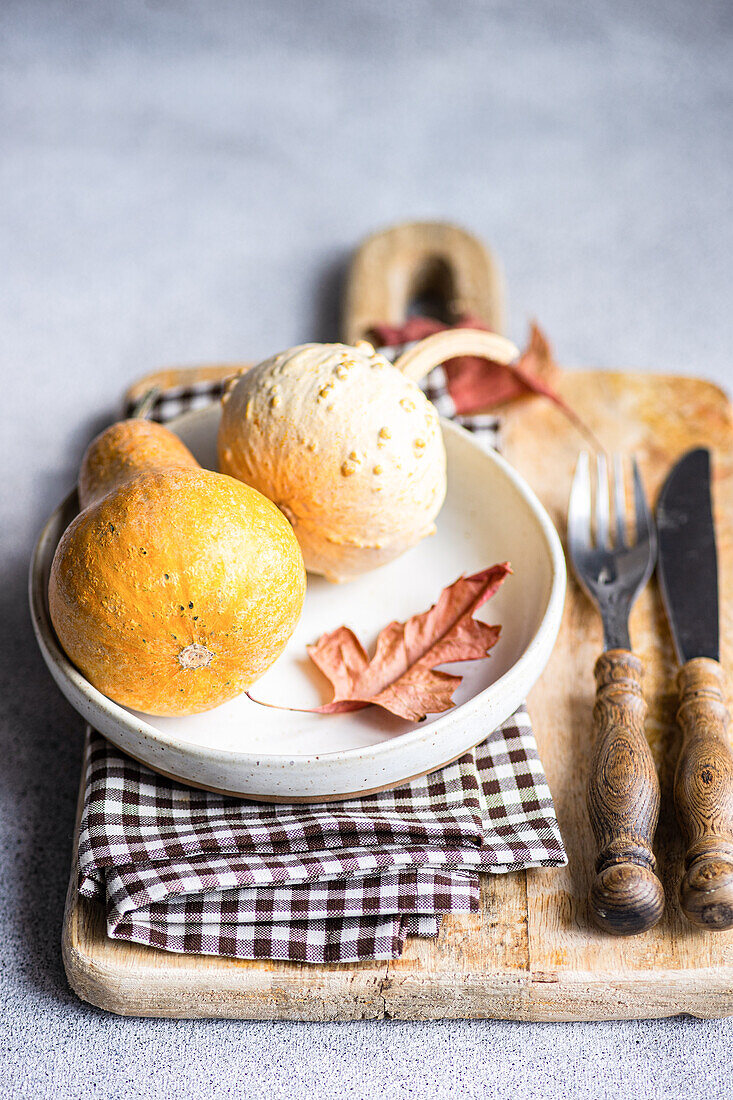 A cozy autumnal flat lay featuring gourds and a dried leaf on a rustic wooden board, accompanied by a checkered napkin and vintage cutlery