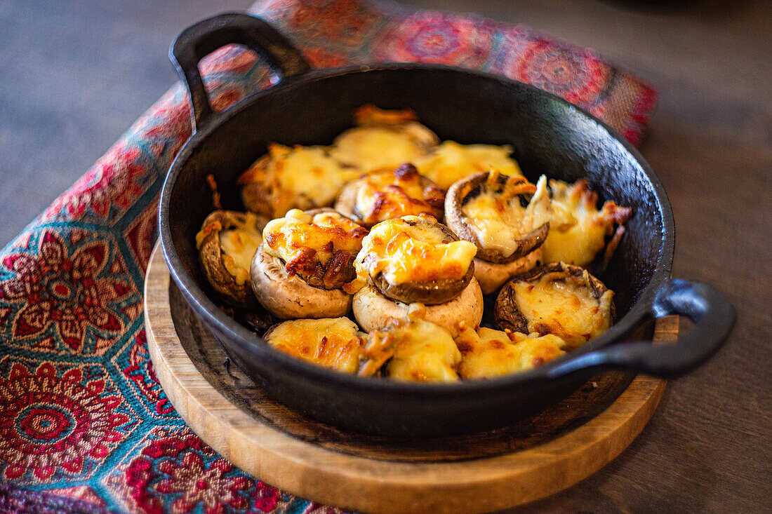 Baked mushrooms stuffed with sulguni cheese served in a black skillet, displayed on a rustic wooden table with a colorful fabric background