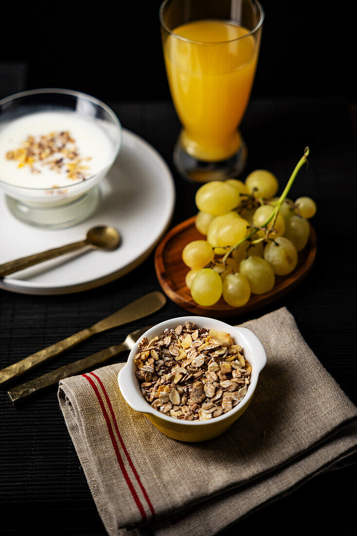 Blick von oben auf ein nahrhaftes Frühstück vor dunklem Hintergrund, mit einer Schüssel Müsli, einem Glas Orangensaft und frischen Weintrauben