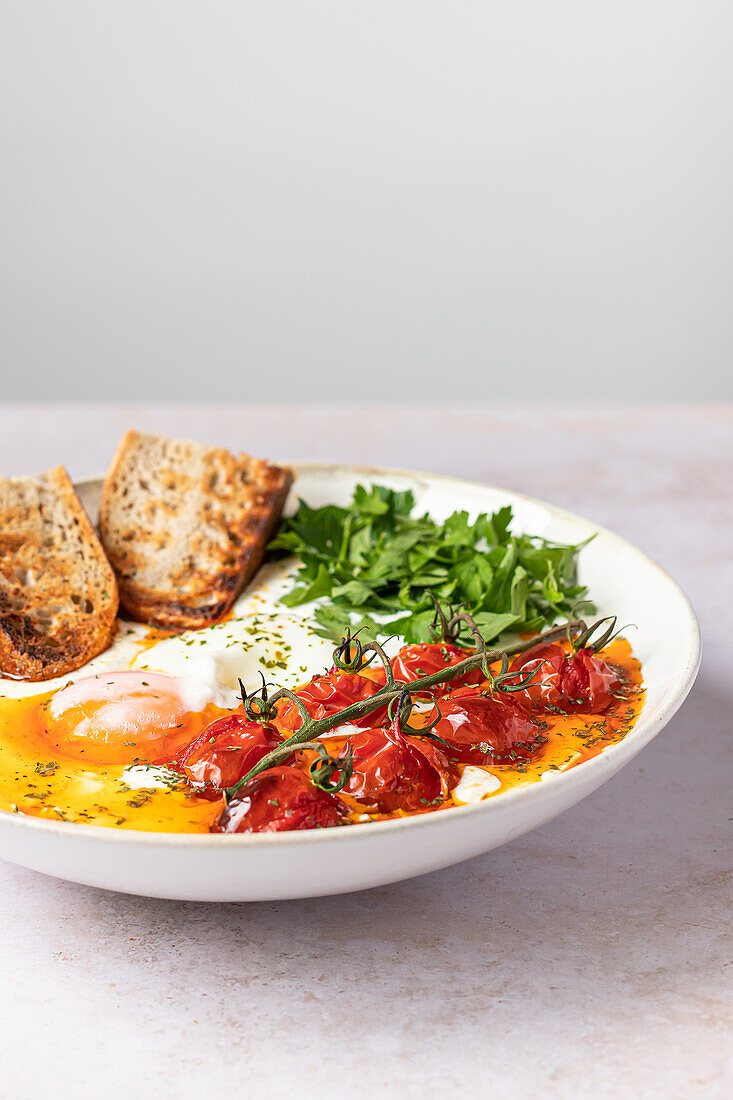 Ein Teller mit türkischen Eiern: pochierte Eier auf griechischem Joghurt, mit gewürzter Buttersauce, Kirschtomaten und Petersilie, dazu geröstetes Brot