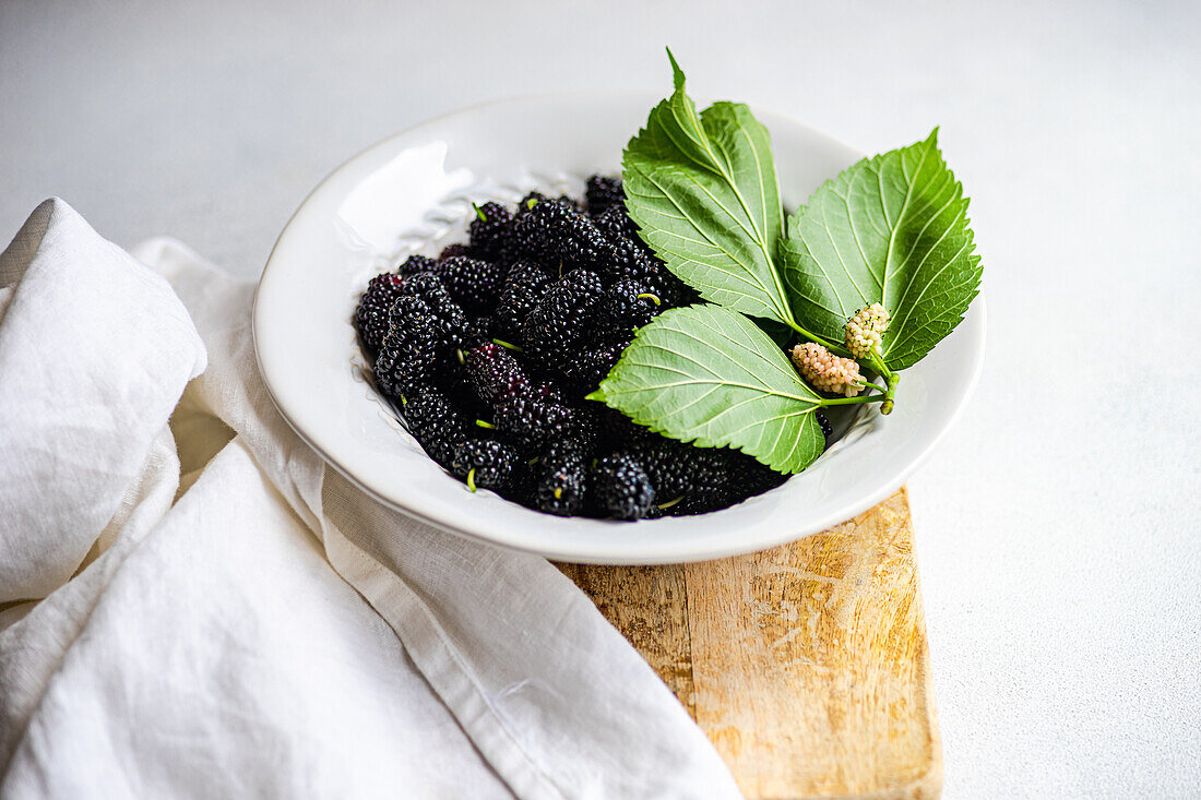 A plate of ripe mulberries accompanied by fresh leaves and tiny unripe fruits sits atop a rustic wooden board, creating a natural and appealing food presentation.