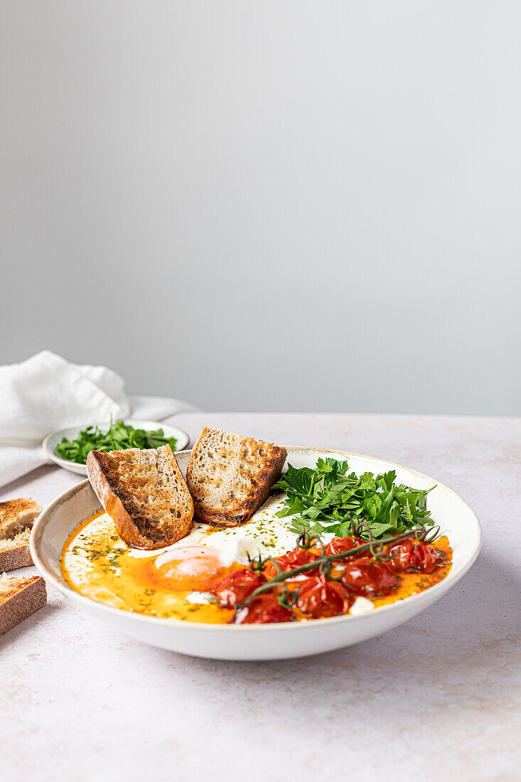 Savory Turkish eggs served on a bed of creamy Greek yogurt, garnished with parsley, alongside seared cherry tomatoes and toasted bread