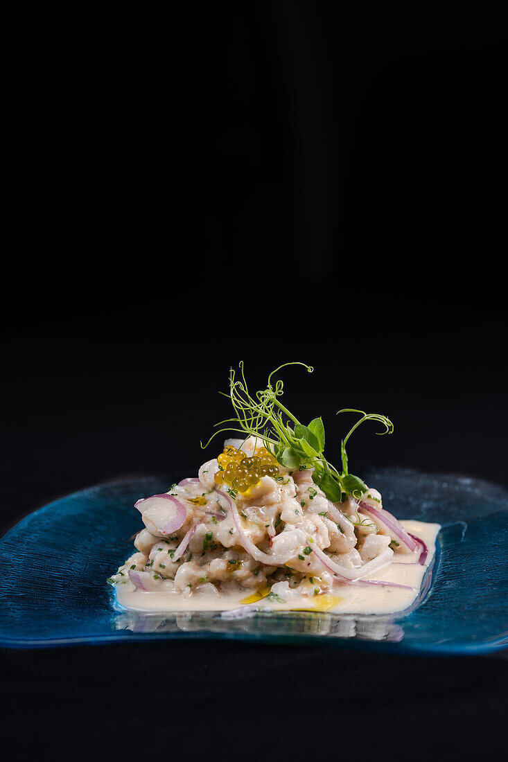 A tantalizing dish of shrimp ceviche garnished with fish roe, sprouts, and red onion, presented elegantly on a blue plate with a dark background.