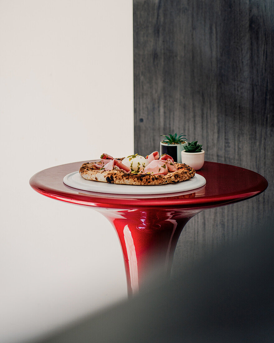 Freshly prepared artisan pizza with toppings and a potted plant on a glossy red table, shot with a soft focus effect