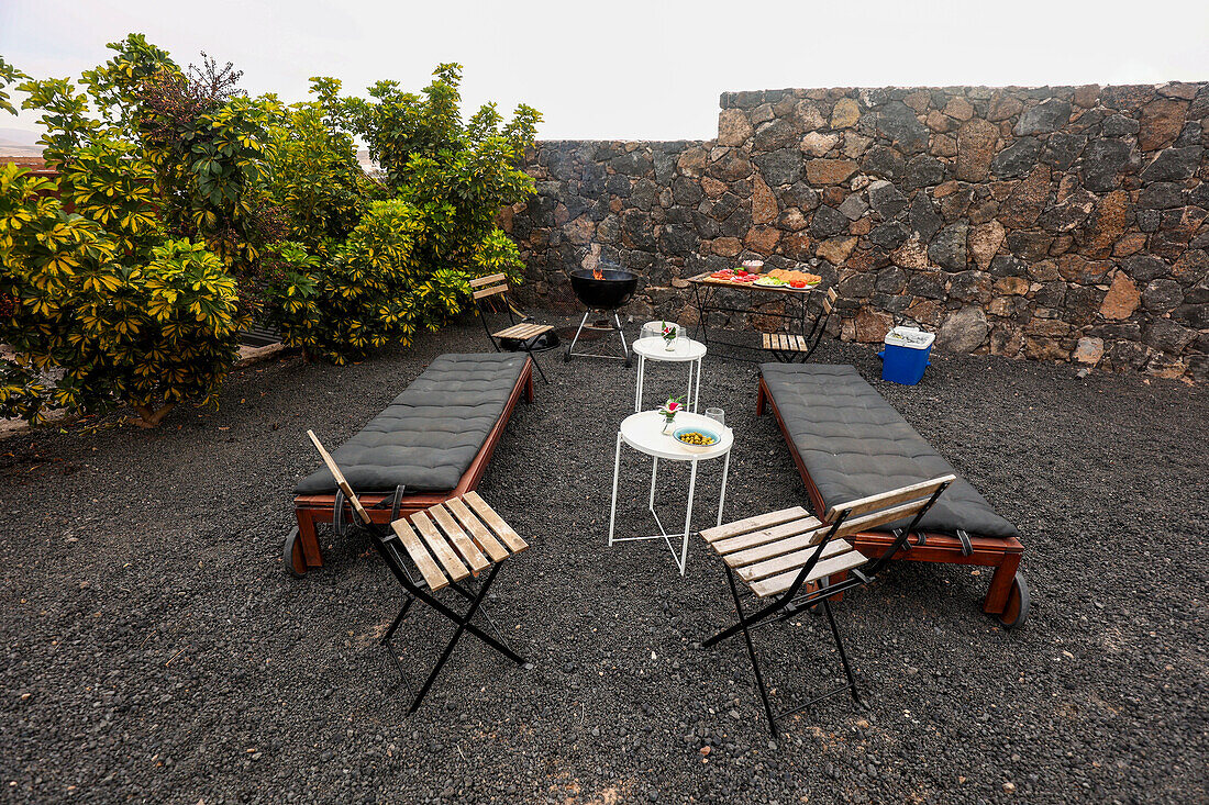 Outdoor area with two lounge chairs, a charcoal barbecue, and three tables displaying food and refreshments readied for a BBQ gathering.