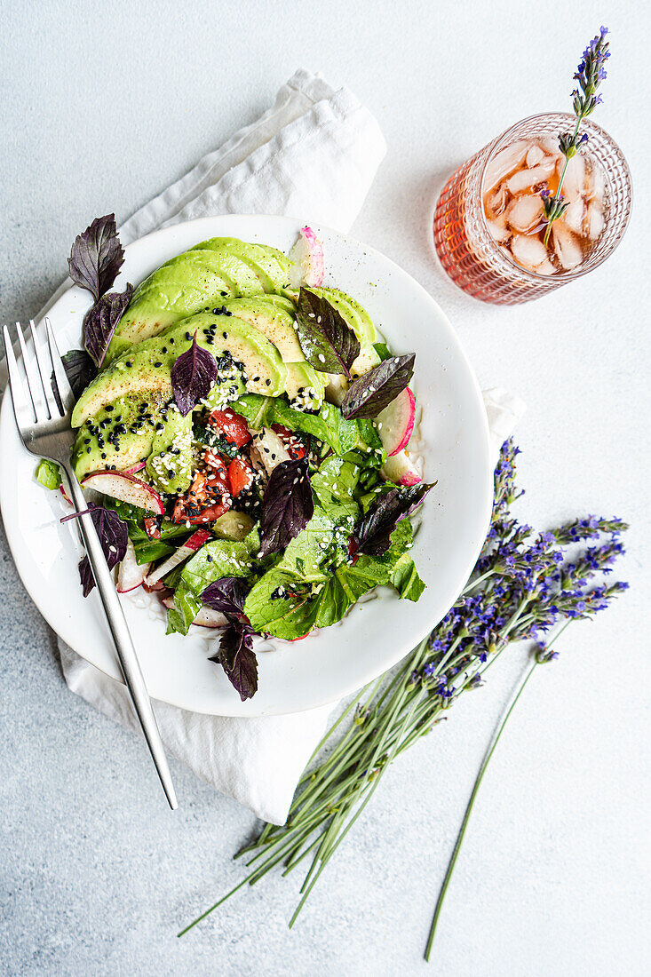 Fresh garden salad with mixed greens, avocado slices, cucumbers, tomatoes, radishes, olives, sesame seeds, and a side of lavender.