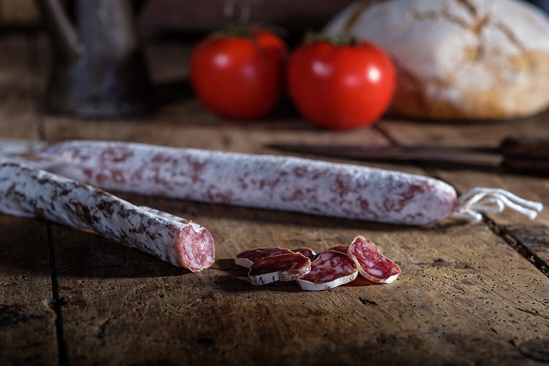 A rustic still life featuring fuet and another type of cured sausage, fresh tomatoes, and crusty bread on an old wooden table, emanating a traditional culinary charm.