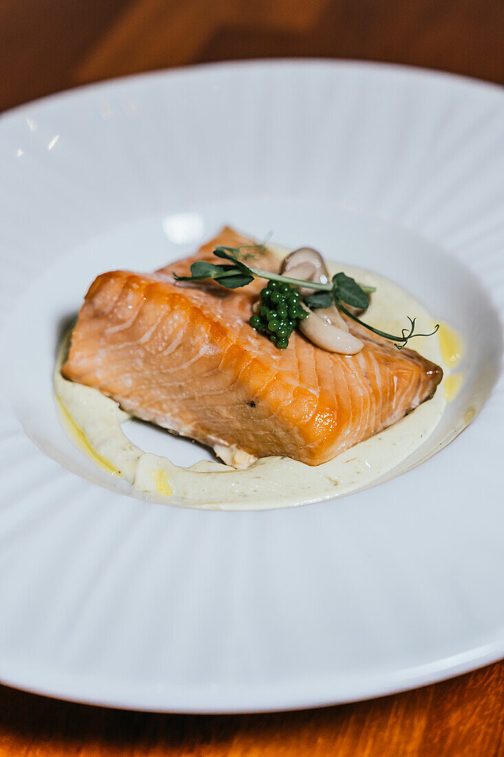 Ein wunderschön pochiertes Lachsfilet ruht auf einer cremigen Unterlage, elegant gekrönt mit Kaviar und frischen Kräutern auf einem weißen Teller