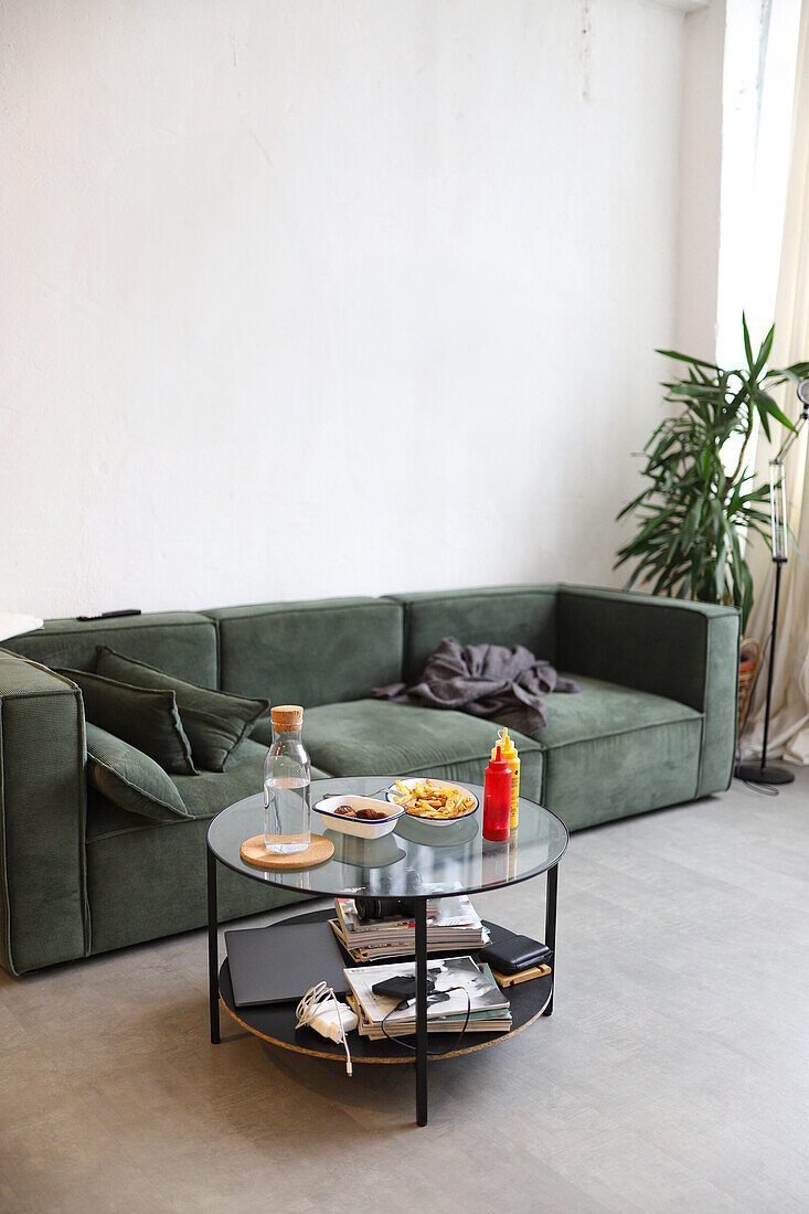 Genießen Sie einen entspannten Tag zu Hause, dargestellt durch ein gemütliches grünes Sofa mit einem runden Couchtisch aus Glas, auf dem eine Reihe von Snacks wie Pommes frites und Soße in einem minimalistischen Interieur stehen.