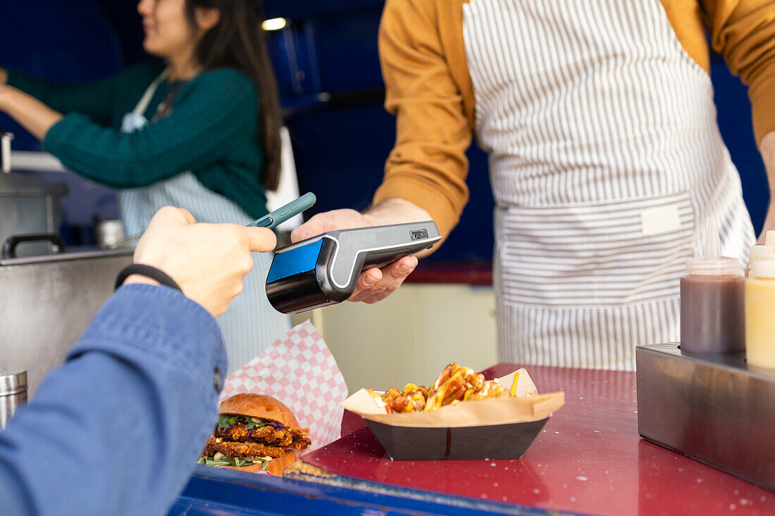 Ein Kunde bezahlt mit einer Karte für Street Food an einem belebten Imbisswagen, während im Hintergrund Freunde zusammenarbeiten