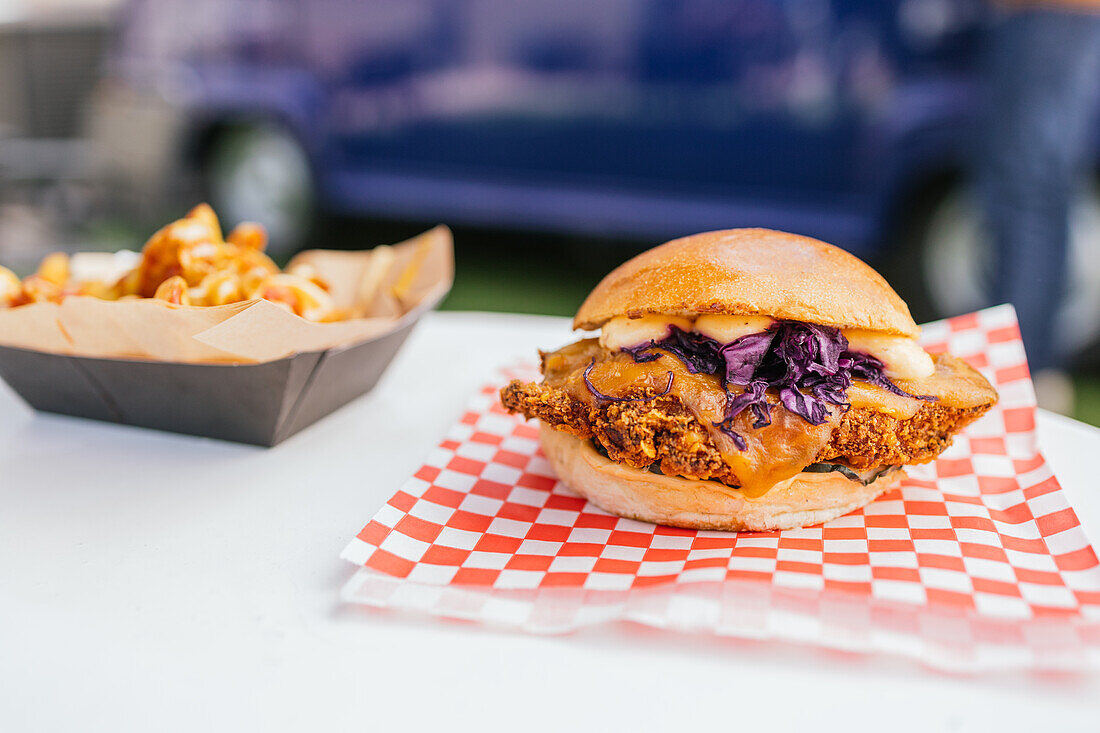 Nahaufnahme eines köstlichen Gourmet-Burgers mit gewürzten Pommes frites, serviert auf einem karierten Papier, mit einem Food Truck im Hintergrund