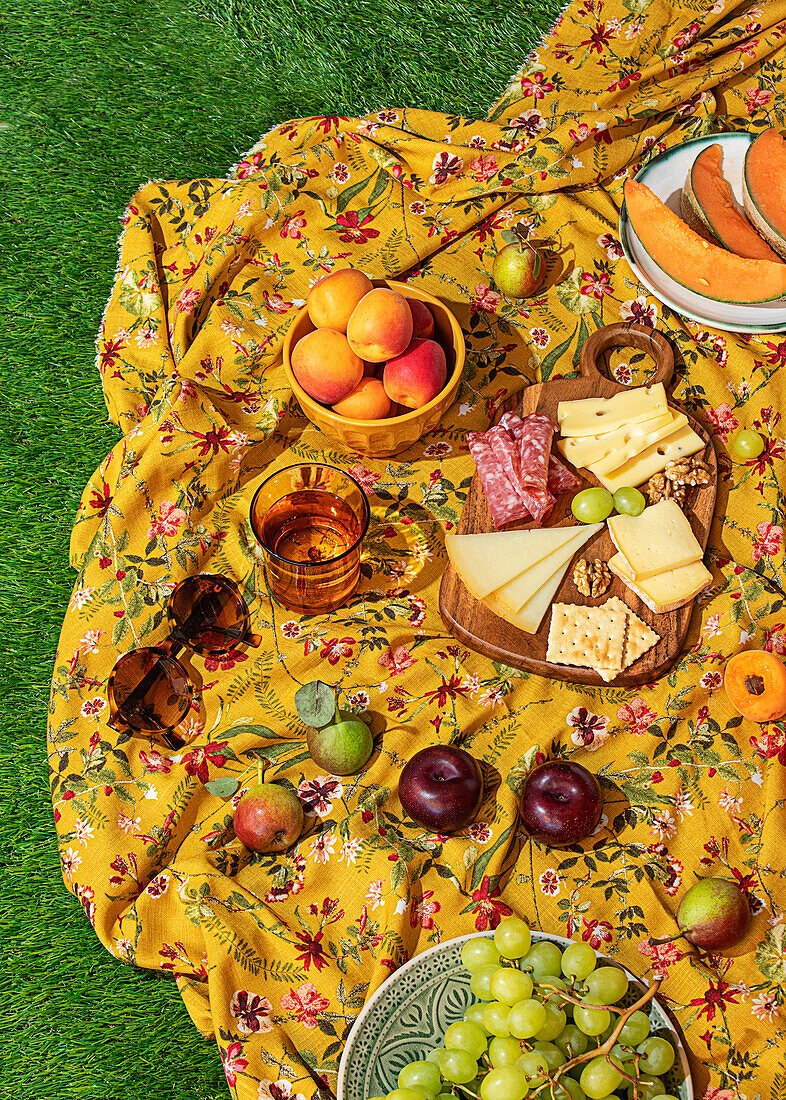 Eine einladende Sommerpicknick-Szene mit einer Vielzahl von Snacks und Früchten, die wunderschön auf einer bunten, blumengemusterten Decke auf üppigem grünen Gras arrangiert sind. Dazu gehören Käse, Fleisch, Weintrauben und Pfirsiche