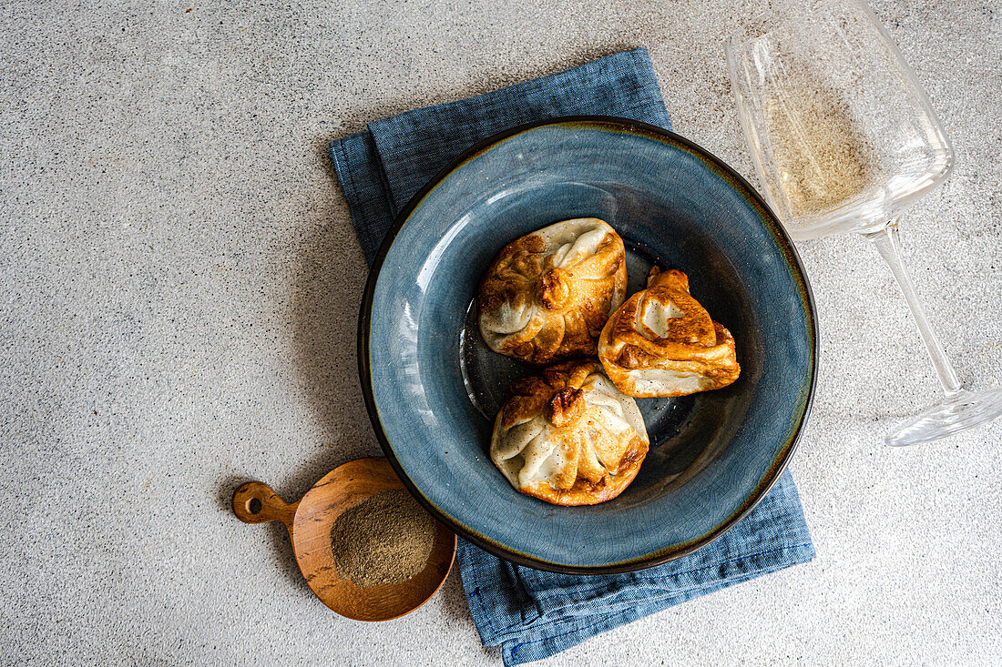 Eine rustikale Präsentation von traditionellem georgischem Khinkali mit goldbraunen Knödeln, die kunstvoll auf einem blauen Keramikteller angerichtet sind. Eine Seite gemahlener Gewürze in einer Holzschale und ein klares Glas liegen im Hintergrund