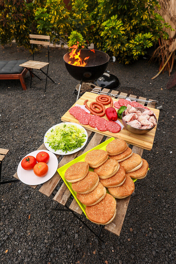 Ein einladender Grillstand im Freien präsentiert eine Vielzahl von rohem Fleisch, darunter Würstchen und Burger, ergänzt durch frisches Gemüse wie Tomaten und Salat, bereit zum Grillen.