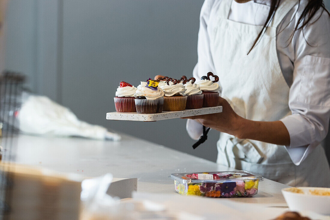 Gesichtslose weibliche Köchin steht mit verschiedenen veganen Cupcakes auf einem Brett in der Küche einer Backstube