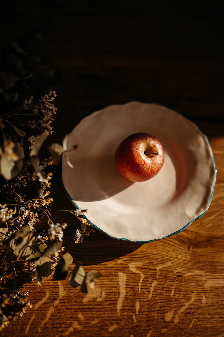 An apple sits in a white bowl on a wooden surface, bathed in warm, dappled sunlight with dried flowers aside