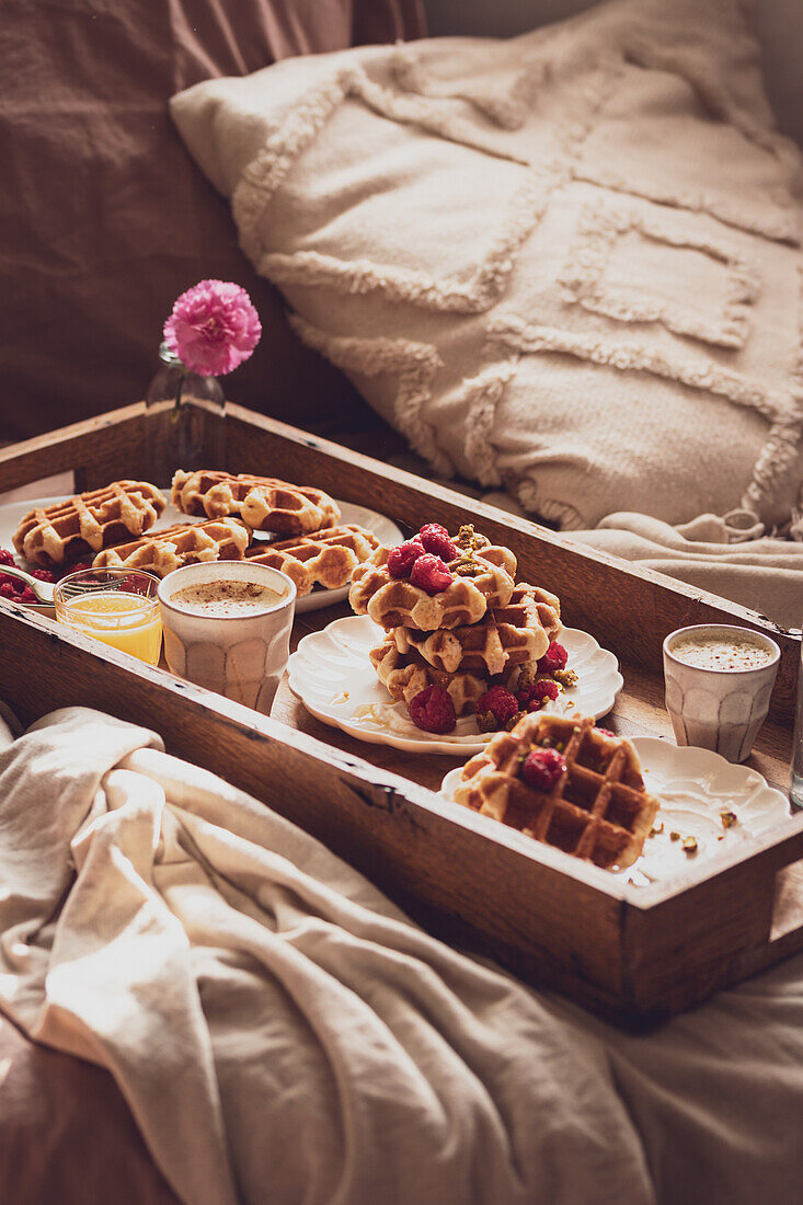 An inviting breakfast tray with freshly baked waffles topped with raspberries, served with coffee and juice, nestled on a snug bed adorned with a cushy pillow and a warm blanket