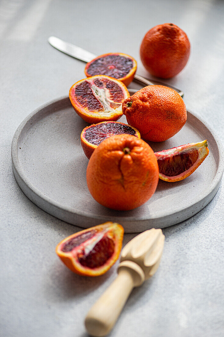 Vibrant blood oranges, both whole and sliced, arranged aesthetically on a gray concrete plate beside a wooden citrus reamer and stainless-steel knife