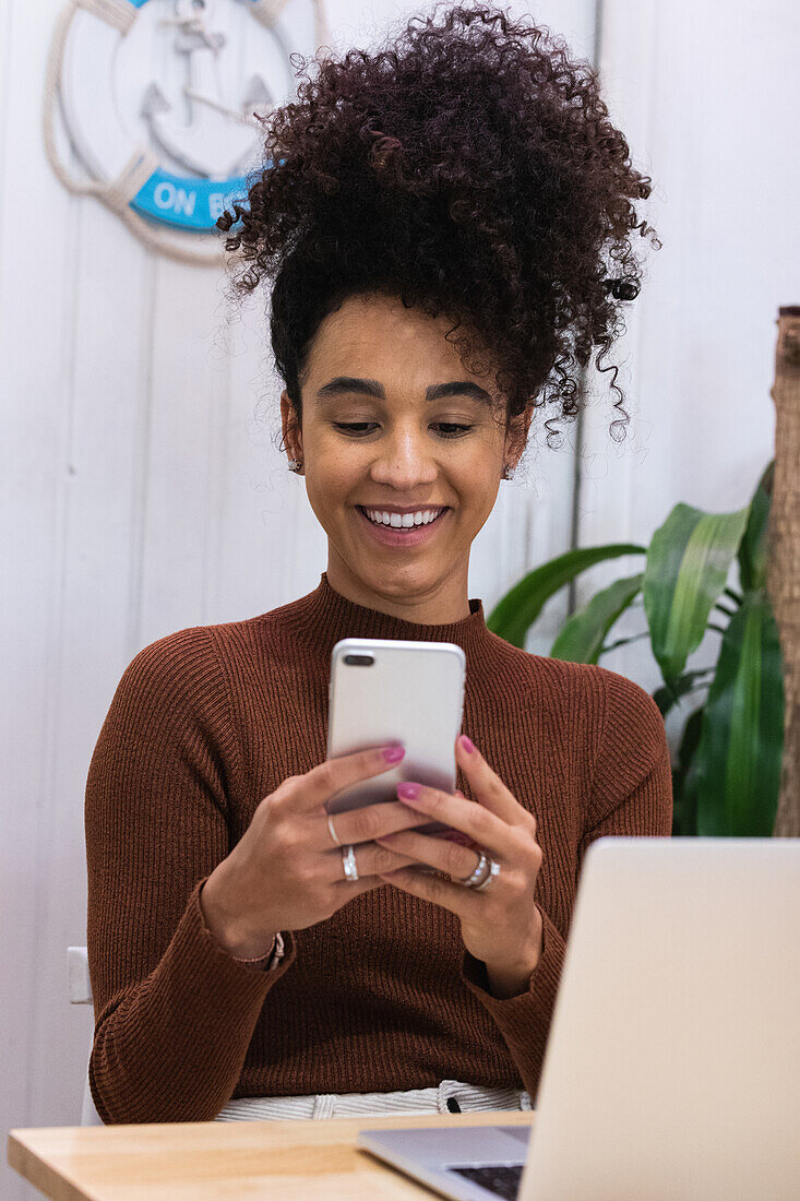 Happy African American female freelancer browsing smartphone and laughing while sitting at table with laptop in cafe and working on remote project