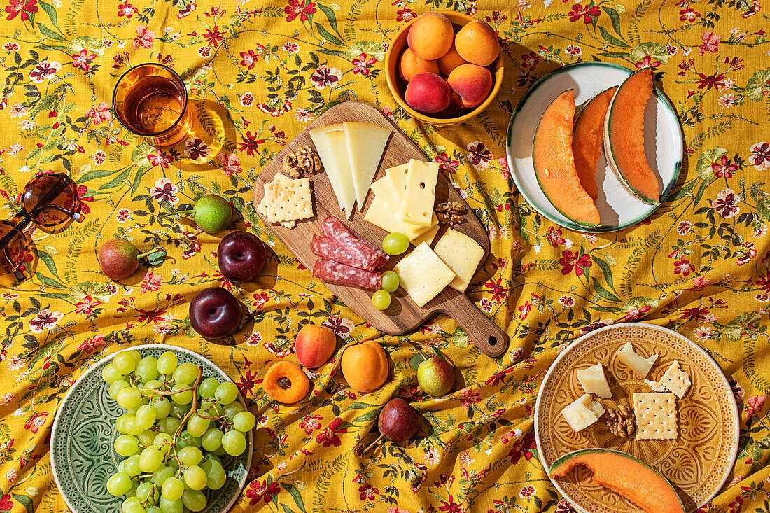 Ein lebhaftes, einladendes Sommerpicknick mit einer Auswahl an Obst, Käse und Wurstwaren auf einer gelben Blumentischdecke.