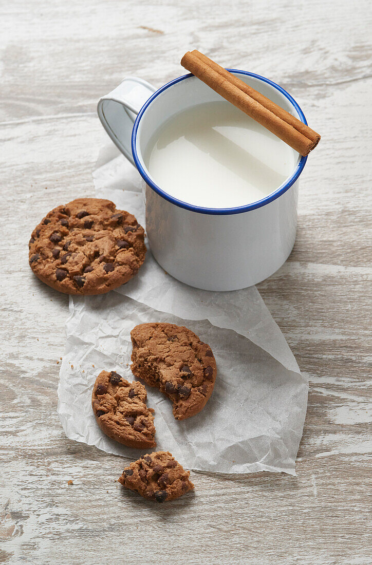 Ein rustikaler Becher Milch, garniert mit einer Zimtstange, neben frisch gebackenen Schokokeksen auf einem verwitterten Holztisch