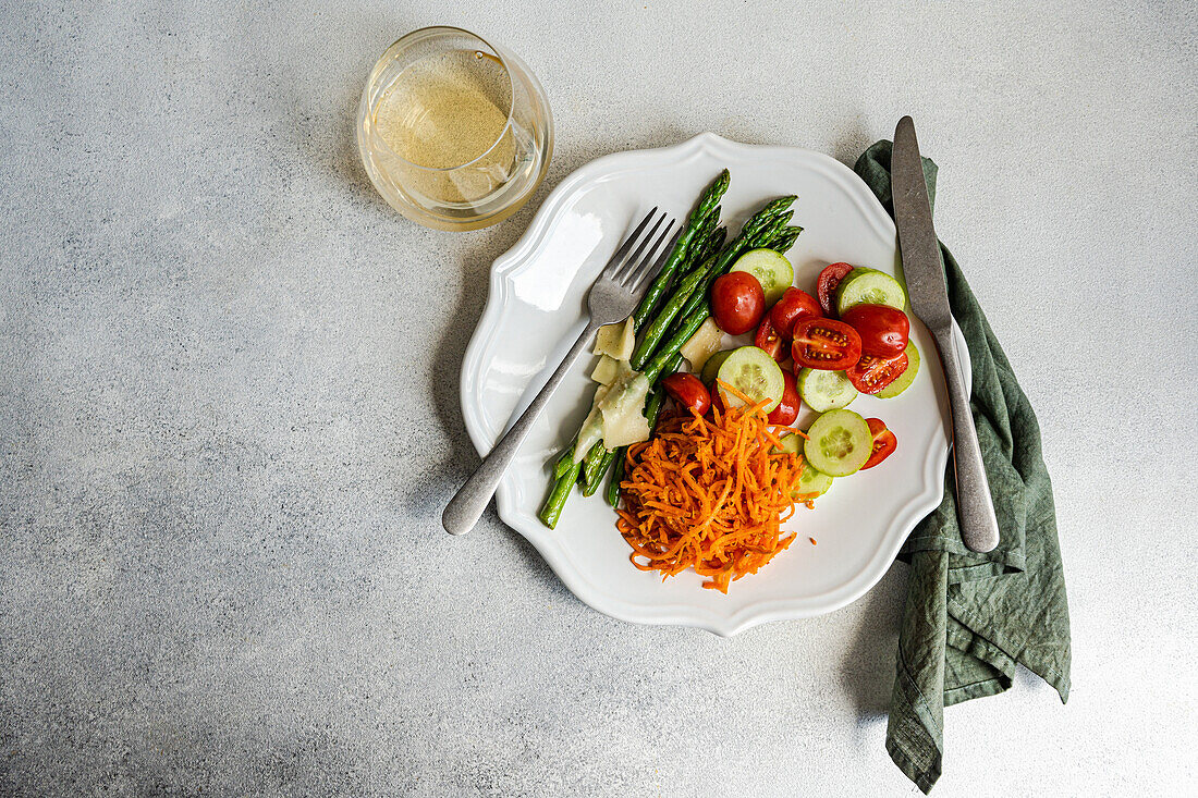 Top view of a healthy, appetizing plate of fresh vegetables featuring steamed asparagus, sliced tomatoes, cucumbers, grated carrots, and cheese, garnished with olive oil and a pinch of salt, served with a glass of wine and a fork on a textured gray background.