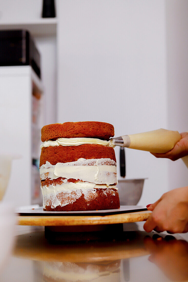 Hands applying frosting on a multi-layer cake with a piping bag, showcasing the baking process