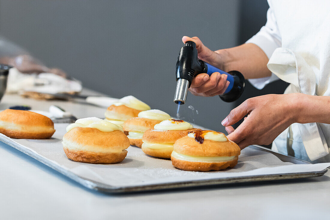 Unbekannter Erntekoch karamellisiert Zuckercreme auf süßen Berlinern mit einer Gasfackel, während er ein veganes Dessert in einer Backstube zubereitet