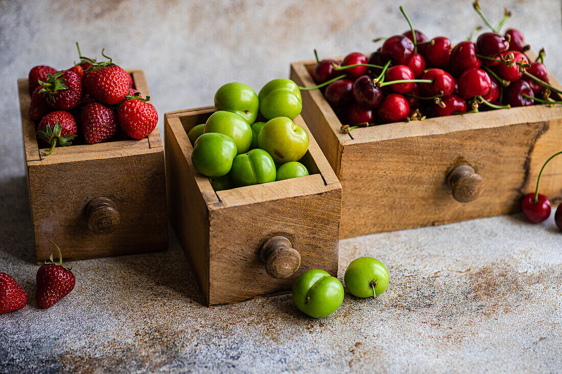 Vintage-Holzkisten, gefüllt mit reifen Bio-Beeren, mit grünen Pflaumen, Süßkirschen und Erdbeeren auf einer strukturierten Oberfläche