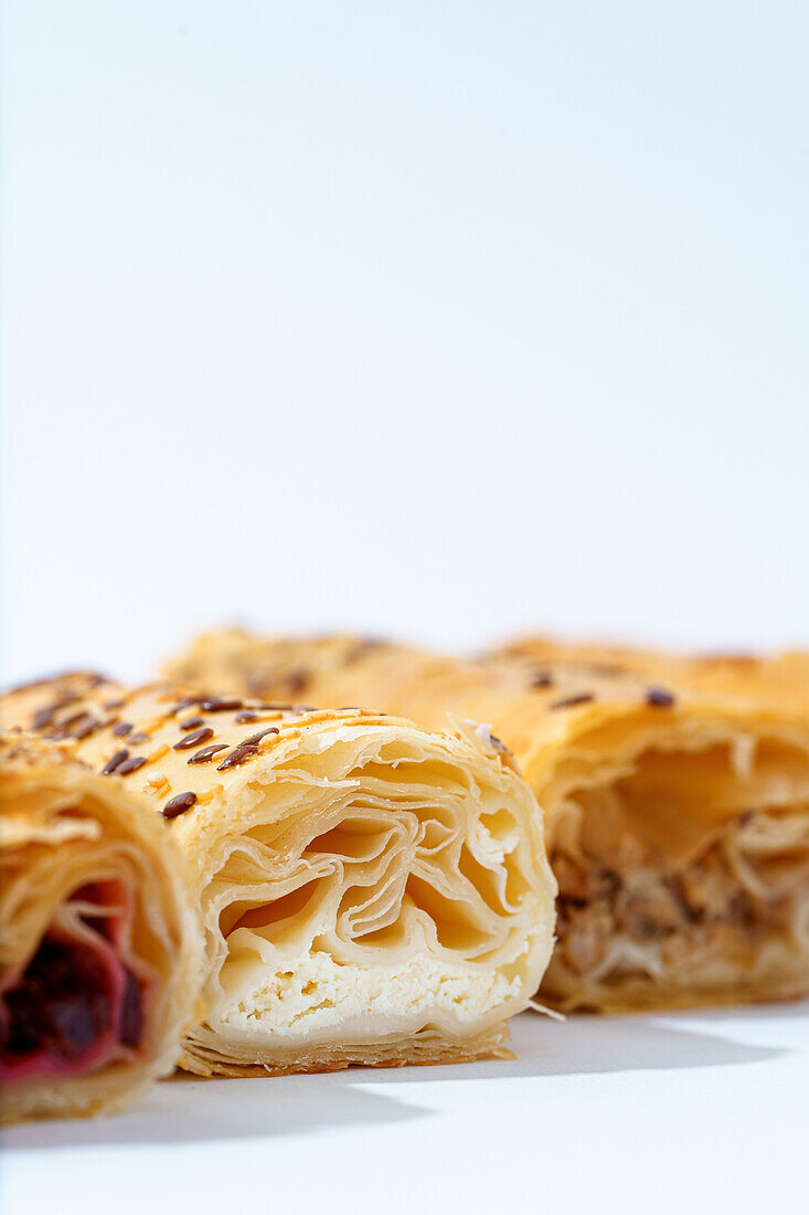 A selection of flaky pastry rolls with various fillings, garnished with seeds, on a clean white background.