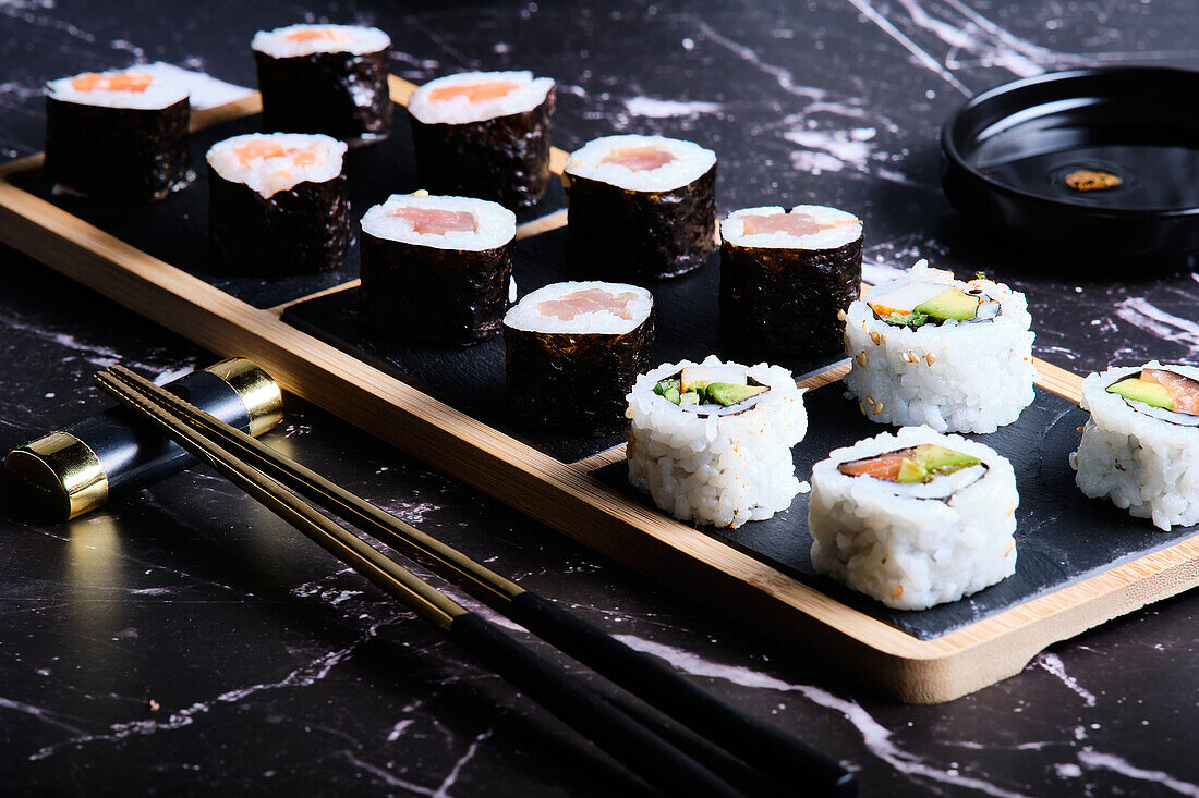 An elegant assortment of sushi rolls elegantly presented on a bamboo plate, complemented by chopsticks and soy sauce on a dark marble background.