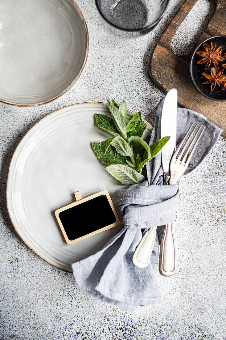 An elegant summer table setting featuring a ceramic plate adorned with fresh mint leaves, complemented by a neat linen napkin and cutlery, set on a textured grey background