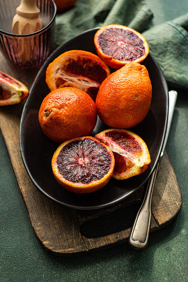 Juicy blood oranges arranged on a black ceramic plate against a rustic wooden board with a green fabric backdrop