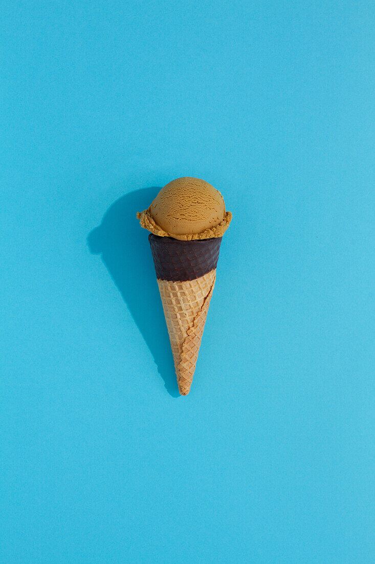 A vibrant coffee ice cream scoop sits atop a classic waffle cone against a blue background, casting a playful shadow